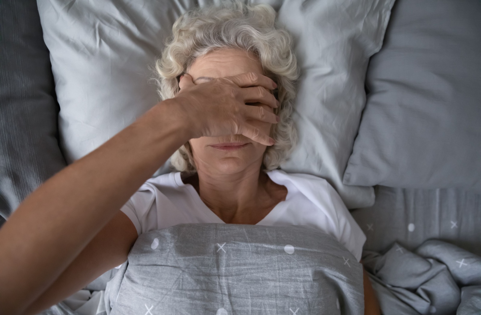 An overhead shot of an older adult lying in bed, rubbing their face in frustration due to trouble sleeping.
