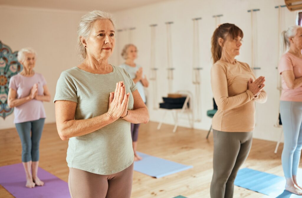 A group of seniors press their palms together while standing on yoga mats, following the direction of a yoga instructor