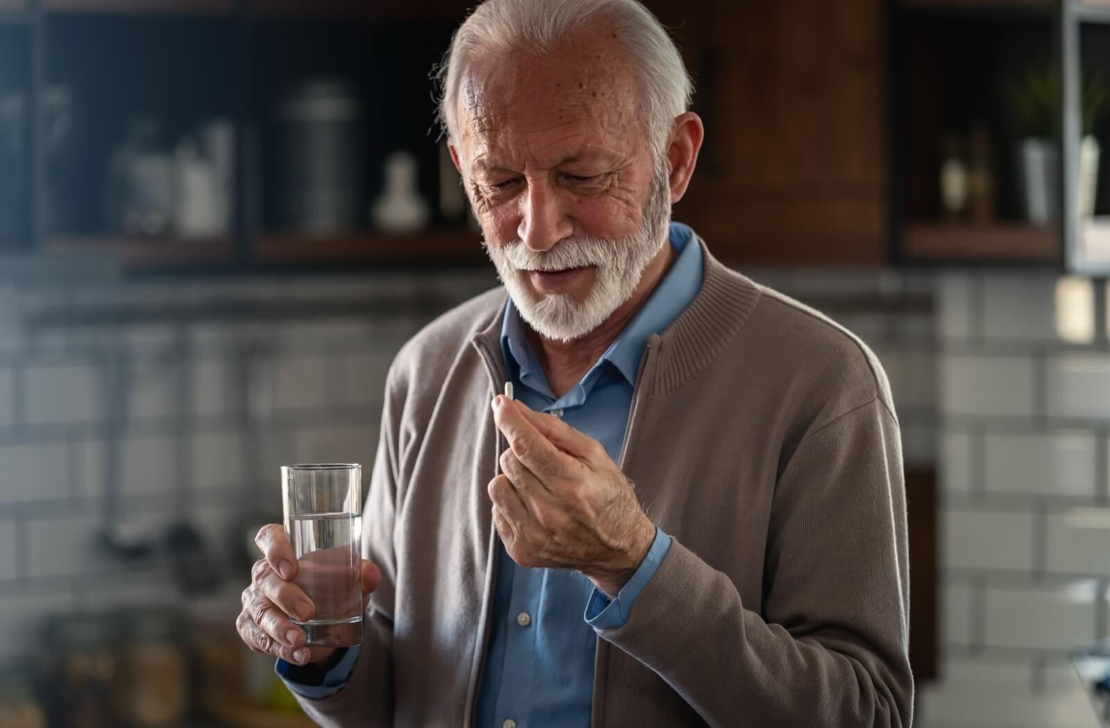 An older adult stands with a glass of water and prepares to take a supplement in their kitchen
