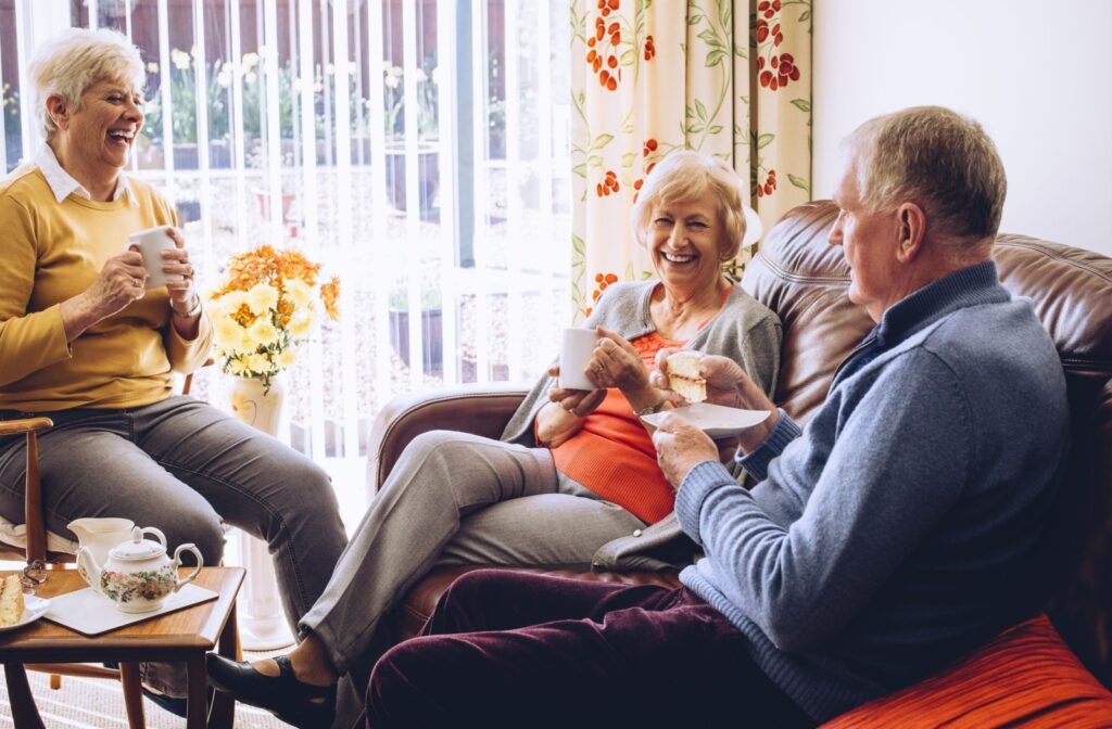 Three older adults sit together, enjoying afternoon tea and snacks, socializing in a cozy assisted living space.