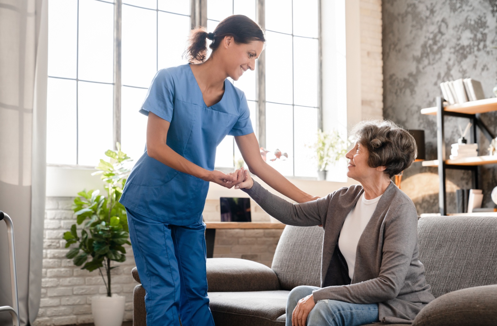 A caregiver in blue scrubs offers support to an older adult in a bright assisted living setting.