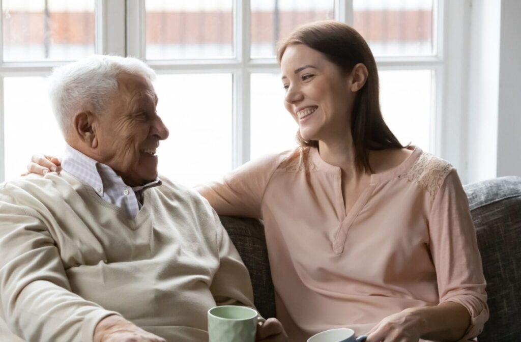 An adult child and their senior parent laugh together and enjoy a warm drink during an assisted living visit.