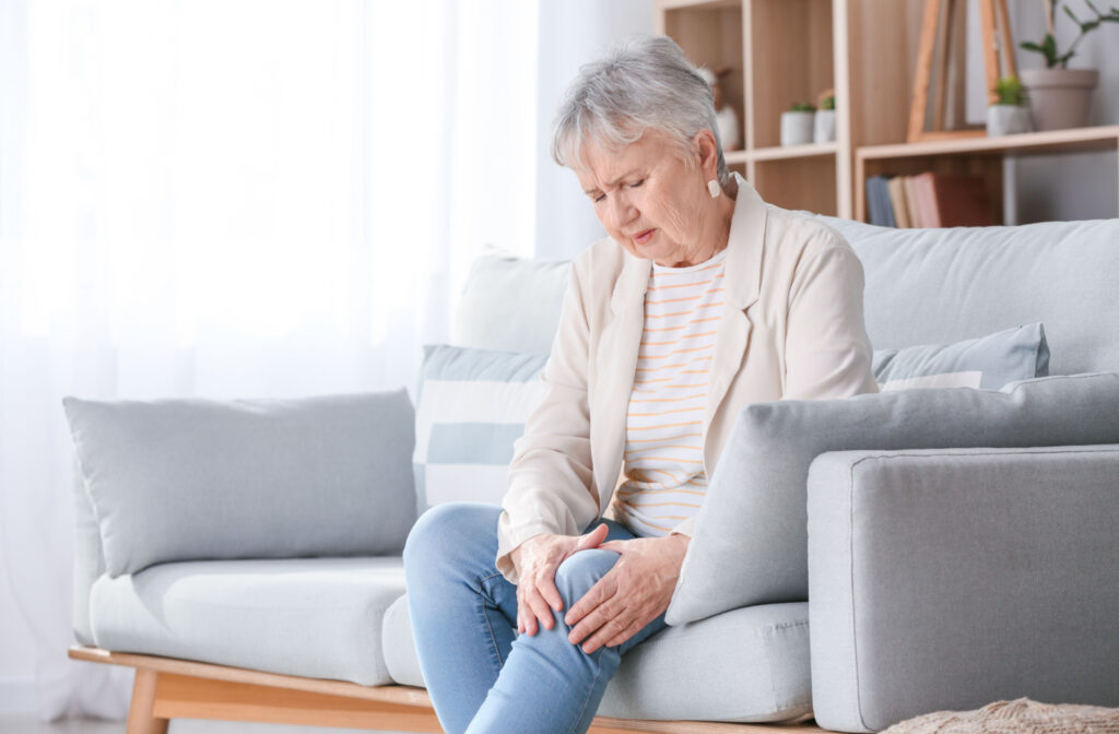 A senior woman sitting on a couch, holding her knee with both hands due to knee pain.