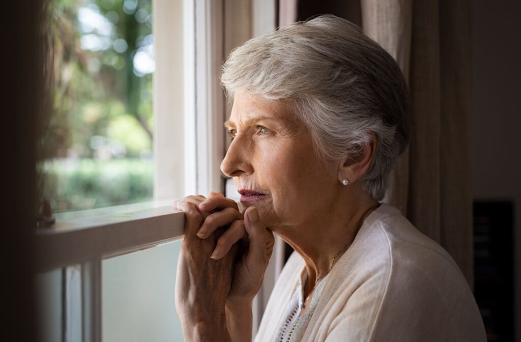 A senior woman holding onto a window sill and looking out of the window with a serious expression.