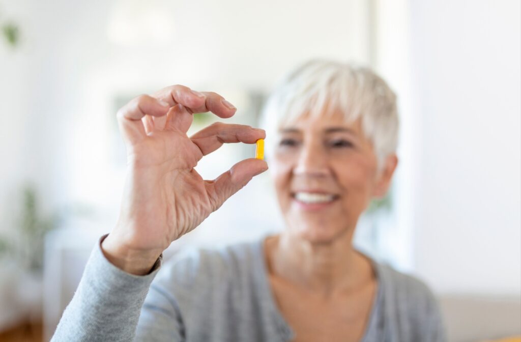 Close-up of a senior woman with short hair holding up a fish oil pill in her right hand against a white background.