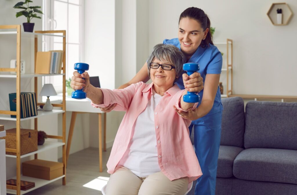 A senior woman in independent living seated and using small weights to regain lost muscle mass while a caregiver supports her.