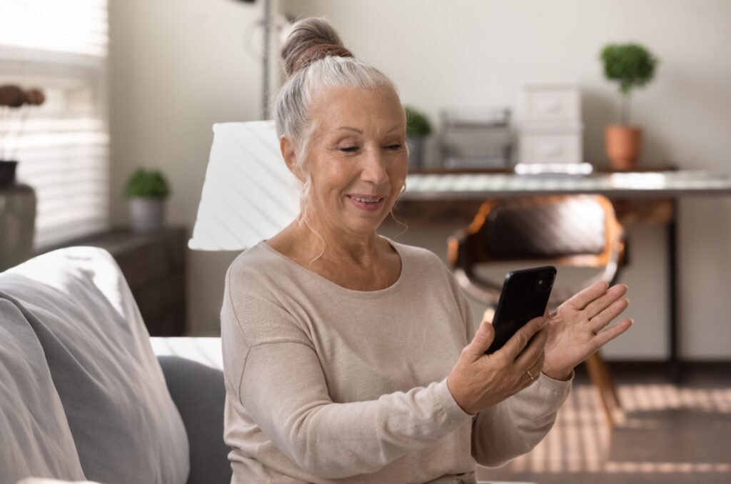 A smiling older adult sitting on a couch talking to family while on a video call.