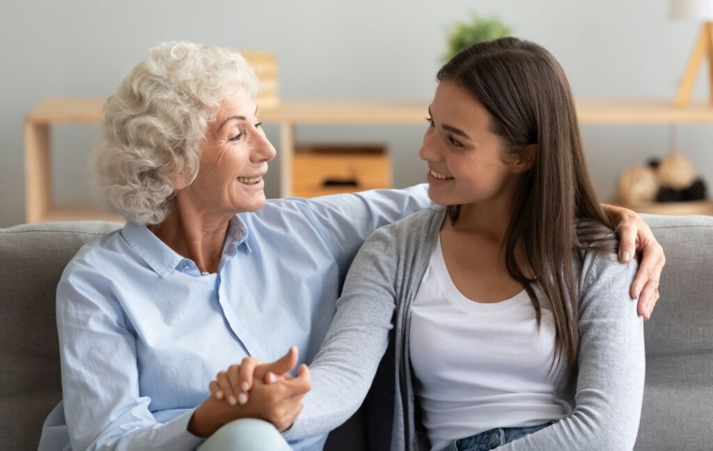 A senior and their adult grandchild are hugging each other as they talk and sit on a couch.