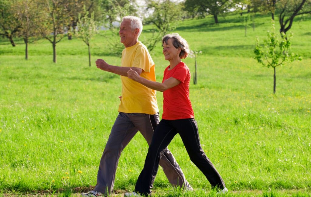 A senior couple wearing active wear are walking briskly though a field of green grass and young trees.