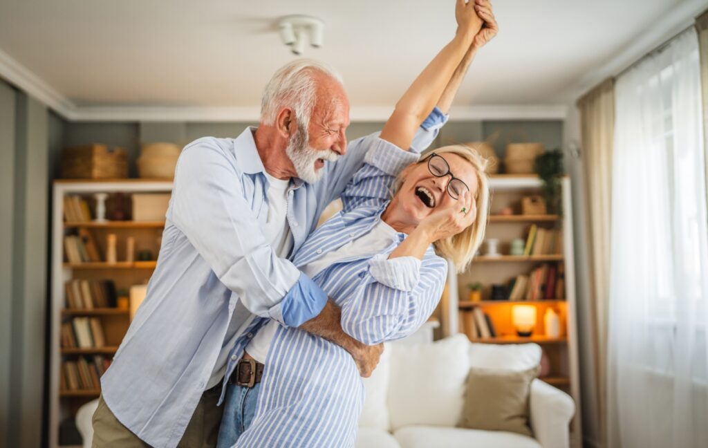 A senior couple are laughing and smiling as they dance together around their cozy living room.