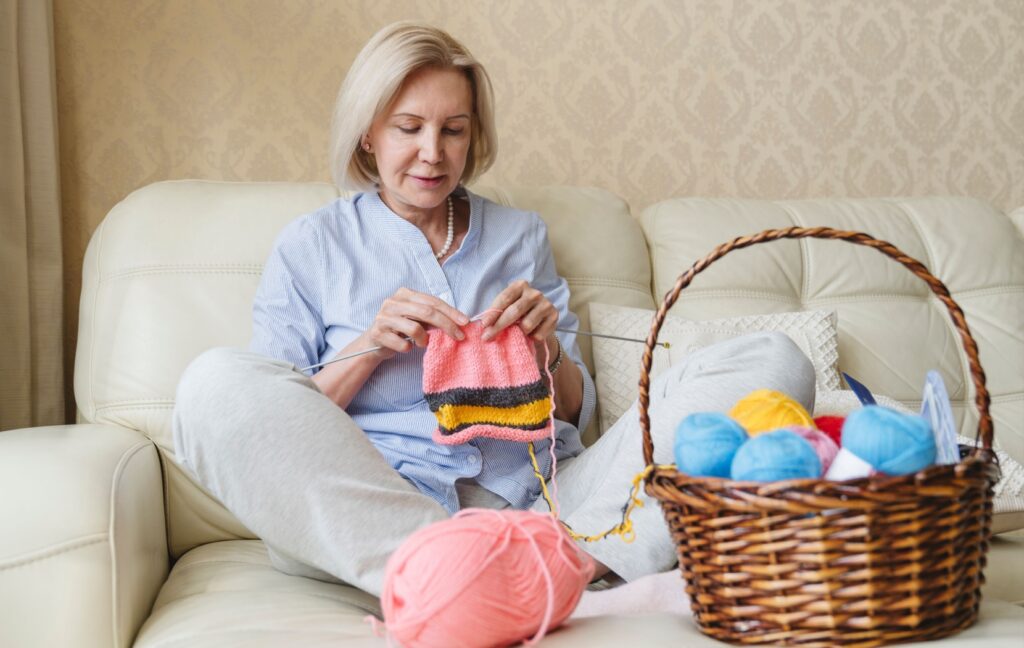 An older adult sits on their cream-colored coach with a basket of yarn and knits a pink scarf.
