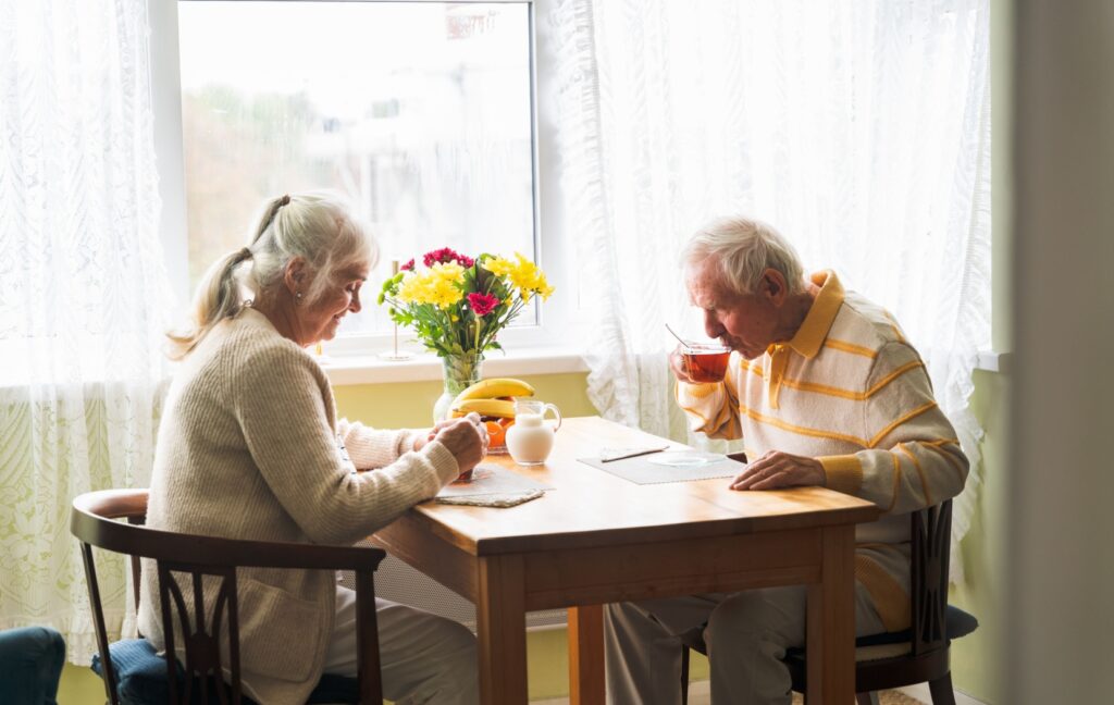 A senior couple eats breakfast in the kitchen of their apartment in an independent living community as the sun shines through the window.