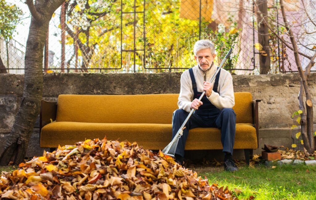 An older adult sits on a patio couch while holding a rake. They stare at a pile of leaves that they need to rake.