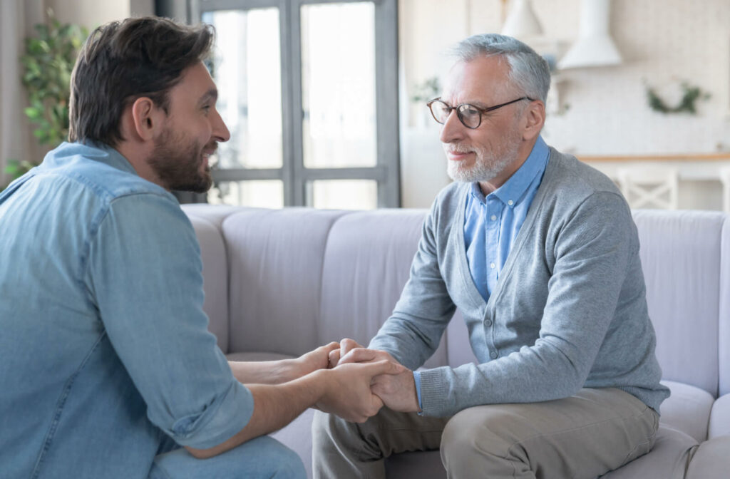 An adult childing holding their older parent's hand and sitting across from them during a conversation about assisted living