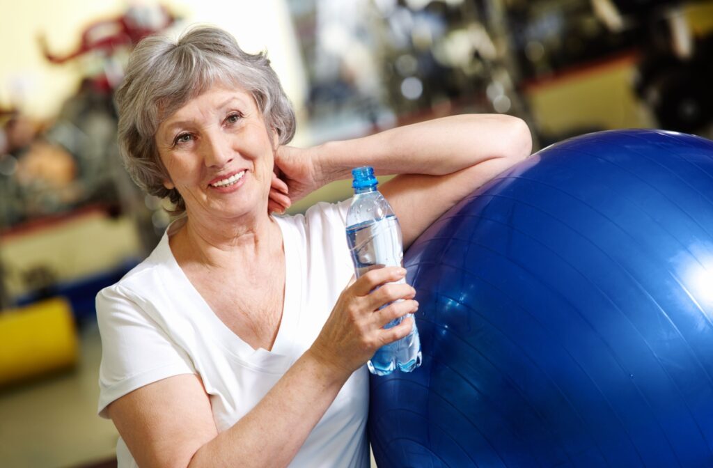 An older adult smiling and leaning on a blue exercise ball while holding an open water bottle.
