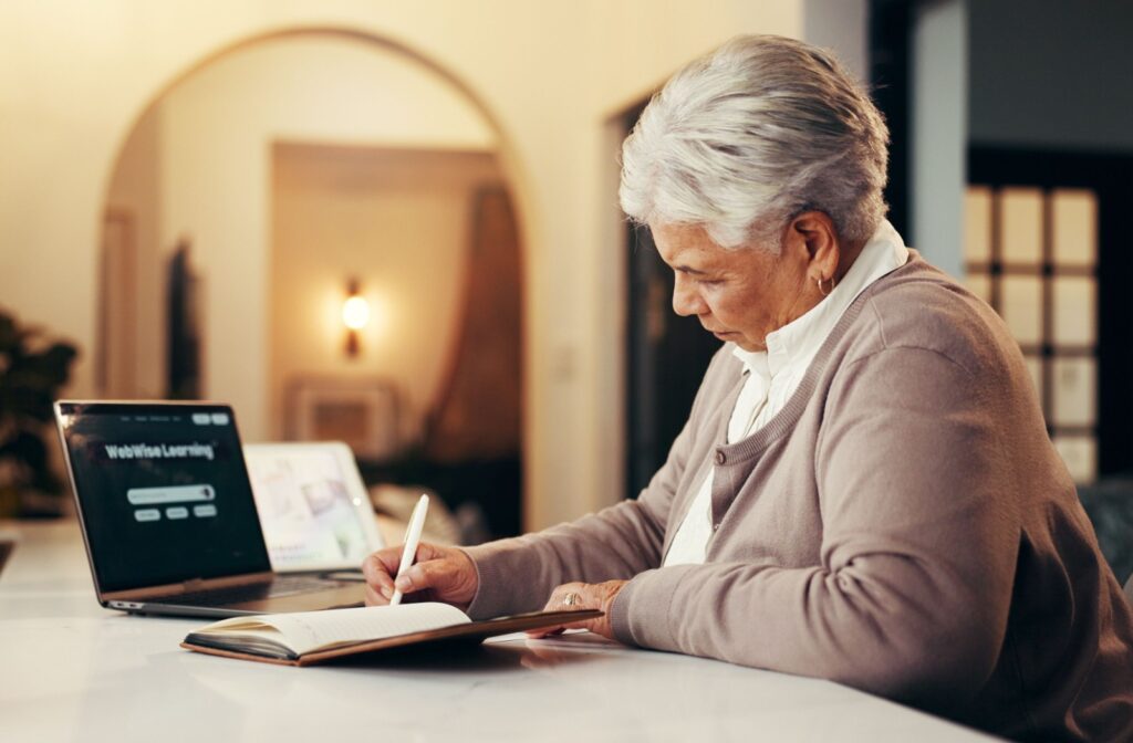 Senior writing notes with laptop open to online learning in a cozy well lit independent living space