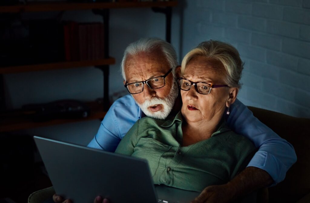 Two older adults sitting close together reacting to something on a laptop in a dimly lit assisted living apartment